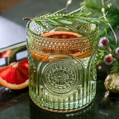 a glass cup filled with liquid next to sliced oranges and greenery on a table