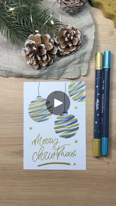 a christmas card next to some pine cones and two pens on top of a wooden table