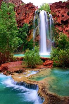 the waterfall is surrounded by green trees and red rock formations, with blue water running down it