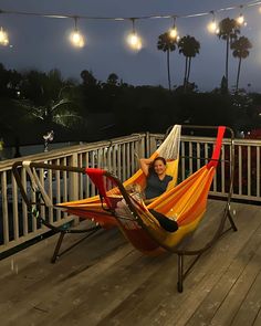 a woman sitting in a hammock on a deck at night with lights strung above her