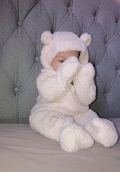 a baby wrapped in a white teddy bear outfit sitting on a bed with his hands to his face