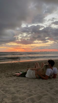 two people sitting on the beach with their backs to each other as the sun sets