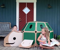 two children sitting on the ground in front of a house made out of cardboard boxes