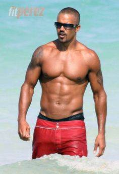 a man with no shirt on standing in the water wearing red trunks and black sunglasses