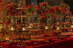 many vases filled with flowers and candles on top of a red cloth covered table