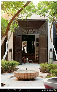 a fountain in the middle of a courtyard surrounded by trees and bushes with an open door