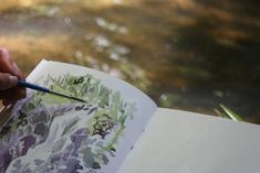 a person holding a pencil and drawing on an open book with water in the background