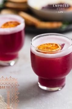 two glasses filled with red liquid and topped with an orange garnish, sitting on a table