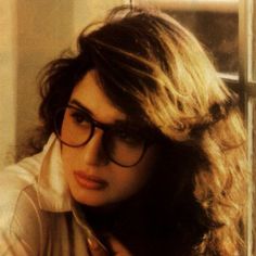 a woman with long hair and glasses leaning against a window sill looking at the camera