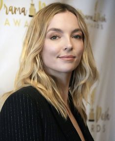 a woman with long blonde hair standing in front of a white backdrop and smiling at the camera