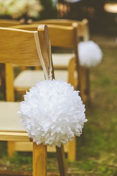 white pom - poms are tied to the back of wooden chairs at an outdoor ceremony