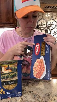 a woman sitting at a kitchen counter reading a box of fustili pasta with an orange and blue hat on her head