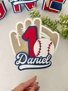 a hand holding up a cake topper with the number one on it and baseballs