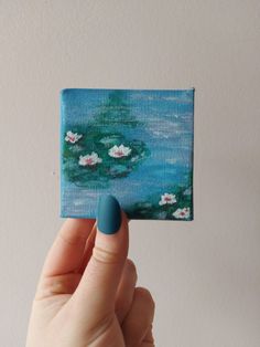 a hand holding up a small square painting with water lilies on the pond in front of it