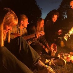 a group of young people sitting around a campfire playing guitars and singing to the music