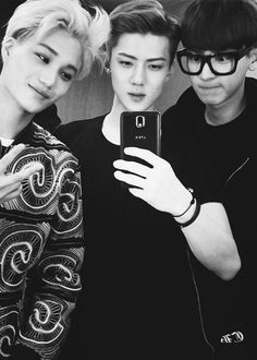 two boys are taking a selfie with their cell phone while another boy is wearing glasses