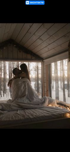 two people sitting on top of a bed in a room with snow covered ground and trees