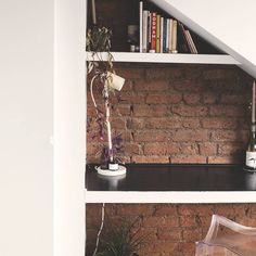 two shelves with plants and books on them in front of a brick wall behind a chair