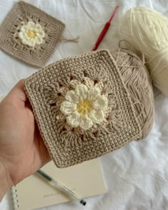 a hand holding a crochet square with a flower on it and two balls of yarn in the background