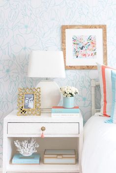 a white nightstand with blue and pink decor on it next to a wallpapered bedroom