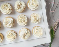 twelve cupcakes with white frosting in a box on a table next to flowers