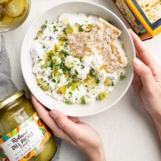 two hands holding a bowl of mashed potatoes with dill pickles on the side