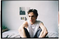 a young man sitting on top of a bed next to a white wall in a bedroom