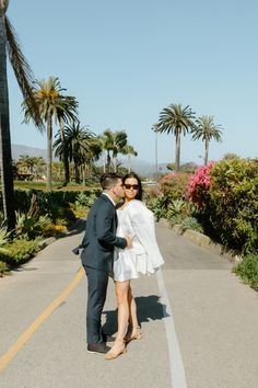 a man and woman standing in the middle of a road with palm trees behind them