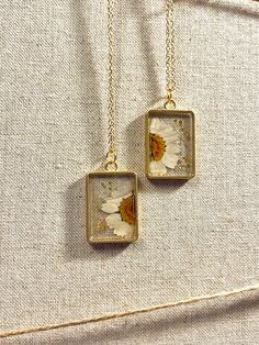 two square pendants with daisies on them are hanging from gold chain necklaces