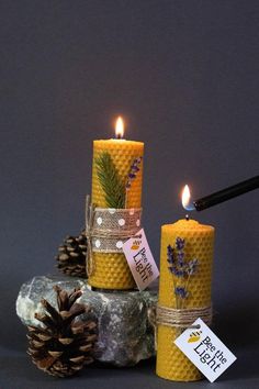 two candles are sitting next to each other on rocks and pine cones with tags around them