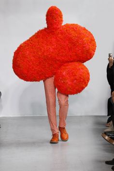 a person with an orange object on their head walking down the runway at a fashion show