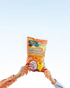 two people holding a bag of popcorn in the air Cpg Food Photography, Popcorn Product Photography, Snack Product Photography, Chips Photoshoot, Popcorn Photoshoot, Evil Photoshoot