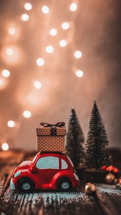 a small red car with a present on it's roof sitting in front of some christmas trees