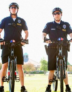 two police officers standing on their bikes in the grass, one is wearing sunglasses and the other wears shorts