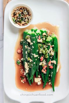 a white plate topped with green vegetables covered in seasoning next to a bowl of sauce