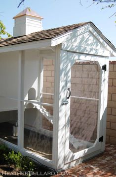 a small white chicken coop with a roof and windows on the top floor, in front of a brick wall