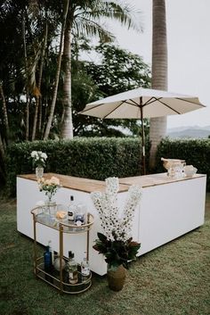 an outdoor bar set up with drinks and flowers on the grass next to it, surrounded by palm trees