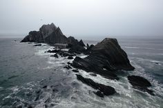 an aerial view of the ocean with rocks in the foreground and foggy sky above