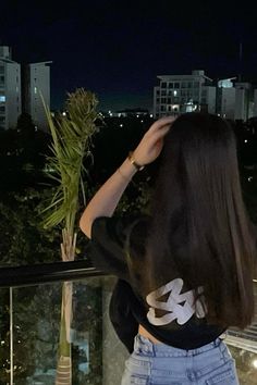 a woman standing on top of a balcony next to a palm tree at night with buildings in the background