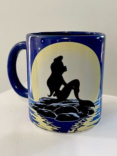 a blue and yellow coffee mug with a silhouette of a mermaid sitting on rocks in the water