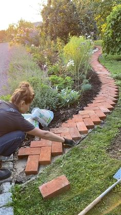 a woman laying bricks on the ground in front of a garden area with flowers and plants