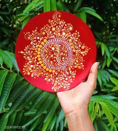 a hand holding a red frisbee with gold designs on it in front of green leaves