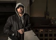 a man wearing a black jacket and hoodie leaning against a wall