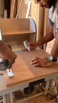 two men working on a wooden table with a cell phone in their hand and another man using a sanding machine