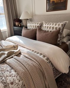 a bed with pillows and blankets on top of it next to a night stand in front of a window