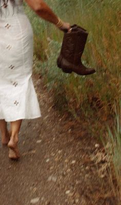 a woman walking down a dirt road holding a brown boot in one hand and a hat in the other