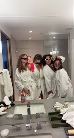 four women in white robes taking a selfie in front of a bathroom mirror and sink