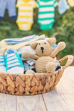 a teddy bear sitting in a basket filled with baby clothes and shoes on a wooden table