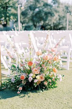 an arrangement of flowers and greenery sits on the grass near rows of white chairs