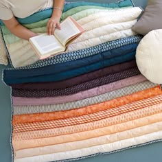 a woman reading a book on top of a bed with pillows and blankets around her